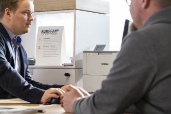To menn sitter ved et bord i en samtale på et kontor med Brother skrivere i bakgrunnen