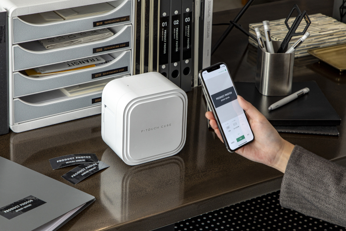 Pöydällä P-touch CUBE PT-P910BT -tarratulostin, johon henkilö tulostaa älypuhelimestaan