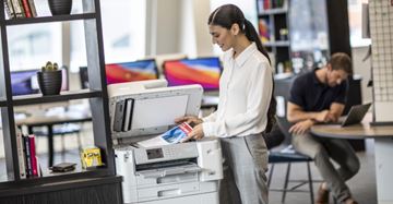 Nainen seisoo toimistossa kopiokoneella ja katsoo tulostamaansa asiakirjaa