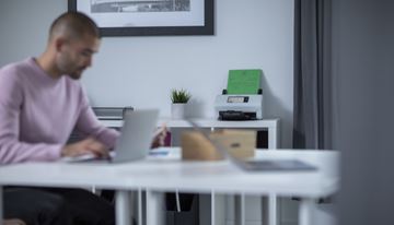 Mies istuu työpöydän ääressä ja tekee työtä kannettavalla tietokoneella taustallaan Brotherin skanneri.