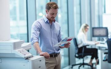 Mies seisoo toimistossa tulostimen vieressä ja katsoo tulostamaansa väritulostetta. Taustalla istuu nainen työpöytänsä ääressä.