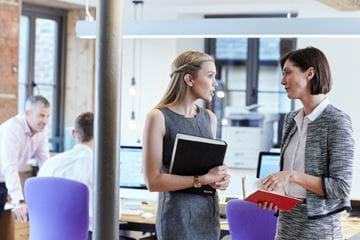Kaksi naista seisovat ja keskustelevat keskenään toimistossa ja kaksi miestä tekevät taustalla töitä yhdessä