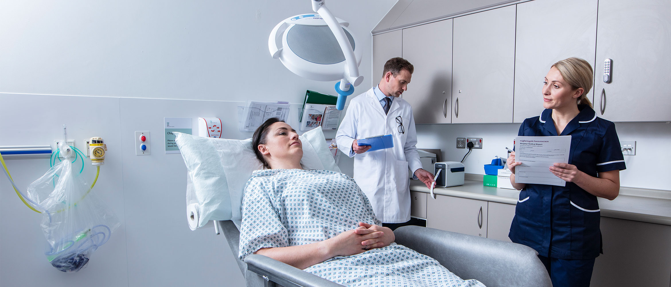 Female patient laid down in treatment room with male doctor in white coat stood at the back and female nurse stood with form