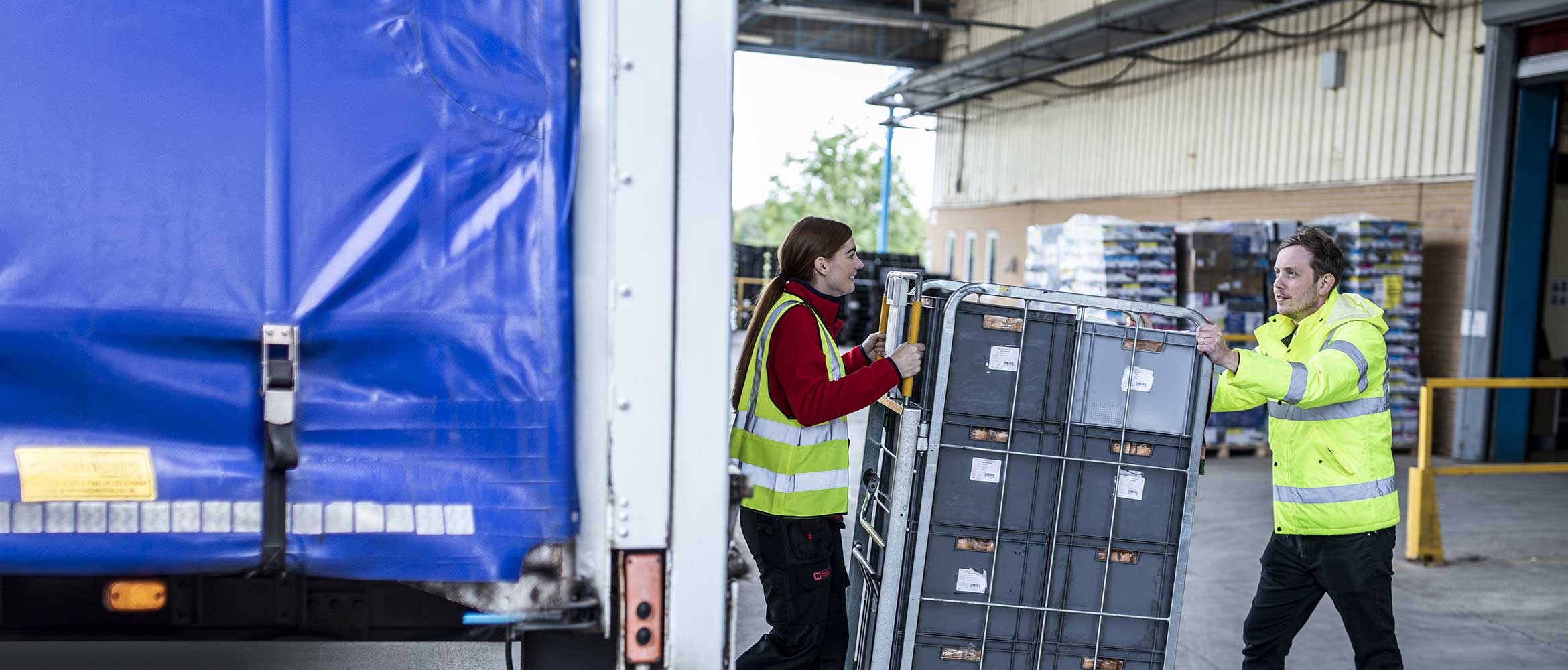 Direct store delivery, two warehouse workers in high visibility jackets moving grey crates onto the back of a lorry