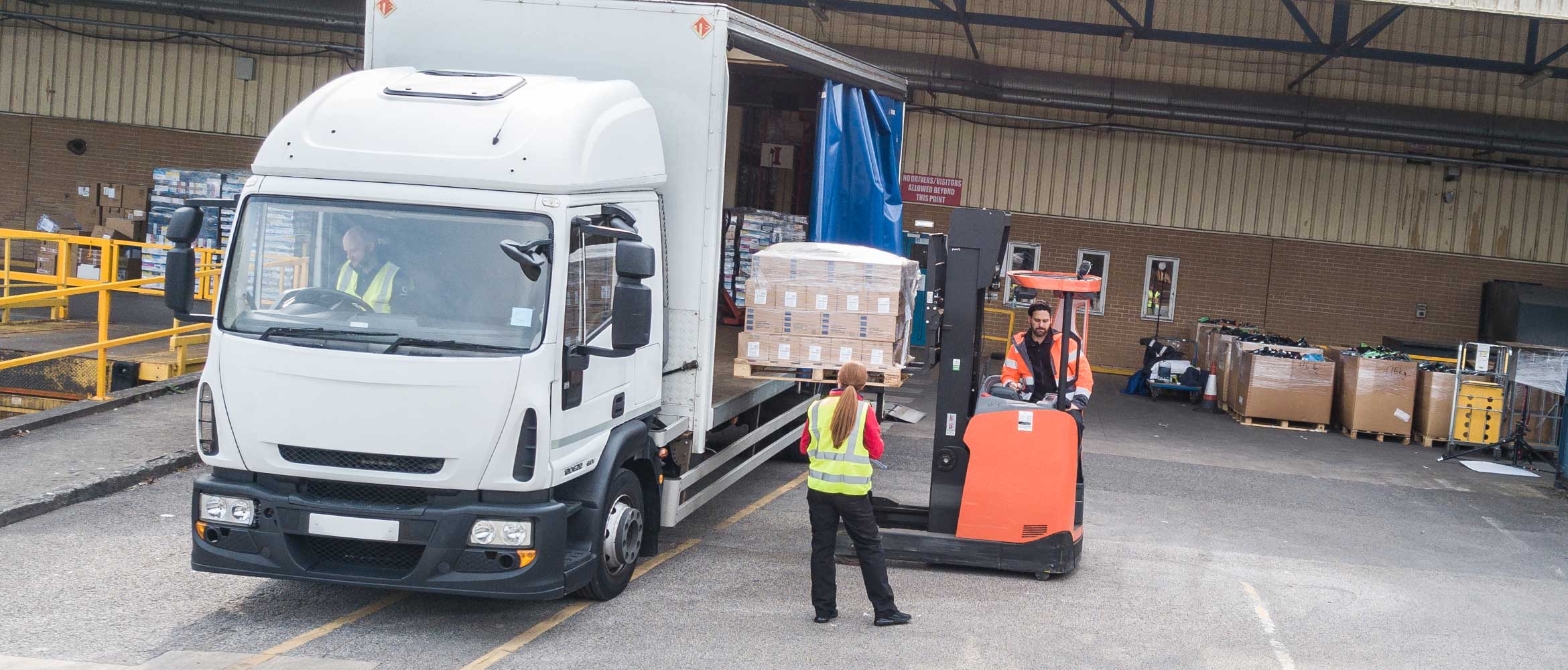 Man sat in truck in-vehicle printing, man in forklift moving pallet into truck, woman stood in hi-vis