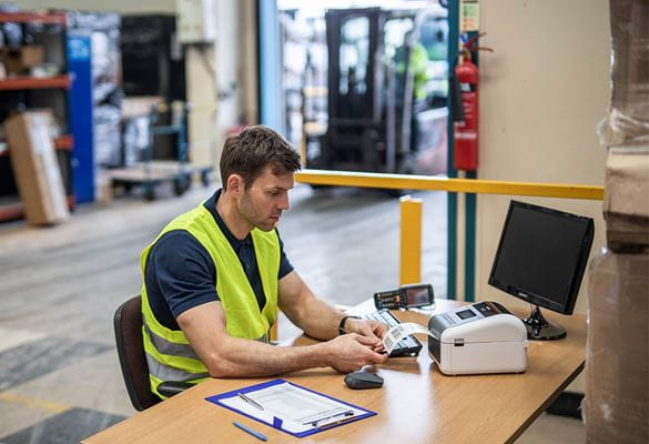 Man with short brown hair wearing yellow hi-vis sat in warehouse at a table, paper, pen, monitor, Brother TD label printer