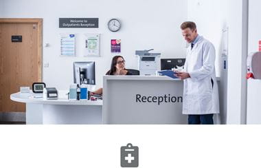 Hospital reception with female receptionist talking to doctor in white coat and grey clip board icon 