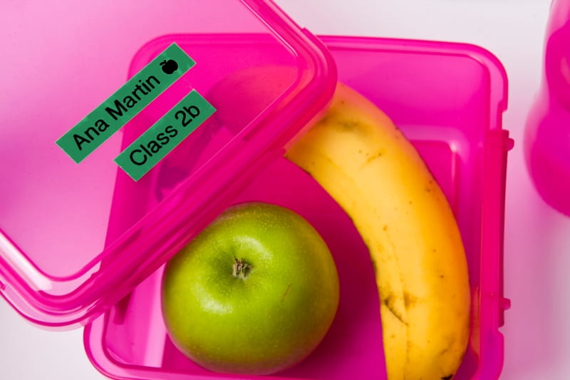 Open pink lunchbox with an apple and a banana inside and a green sticker label on top
