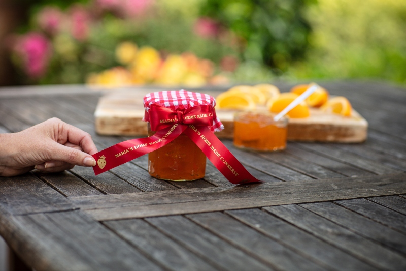 Hand holding a red printed ribbon tied to a jam jar, on a table with some sliced oranges in the background