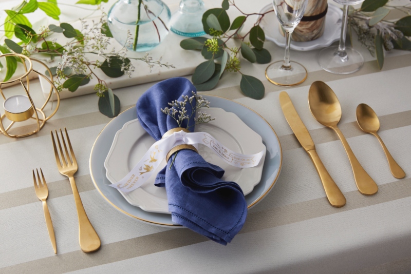Table setting with gold cutlery, glasses and white plate with a blur napkin tied with a white printed ribbon
