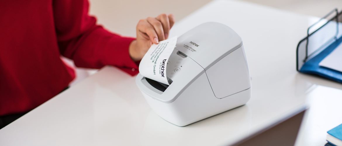 A lady wearing a red top removing a printed address label from a Brother thermal label printer which is on a desk in an office environment
