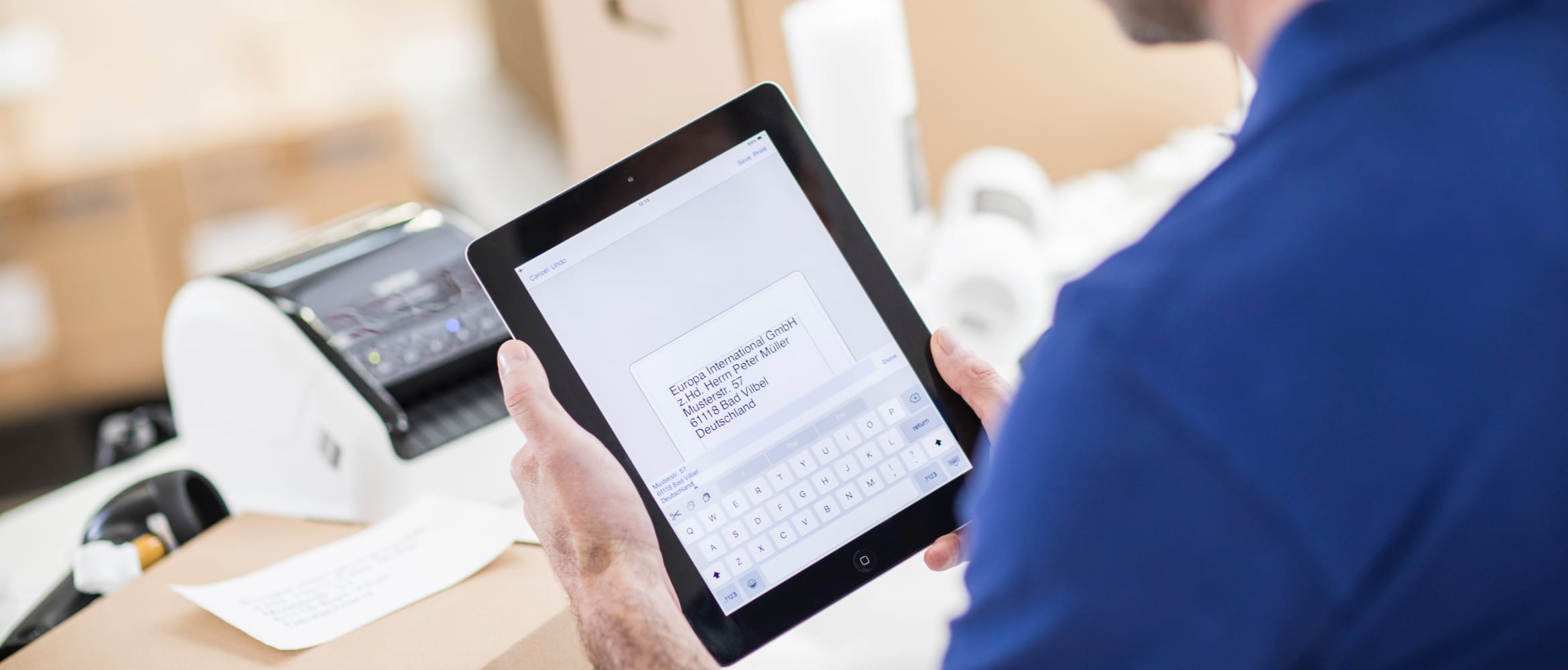 A man using Brother's iPrint&Label app on a tablet device to print a return shipping label on a Brother label printer, with packing boxes in the background.