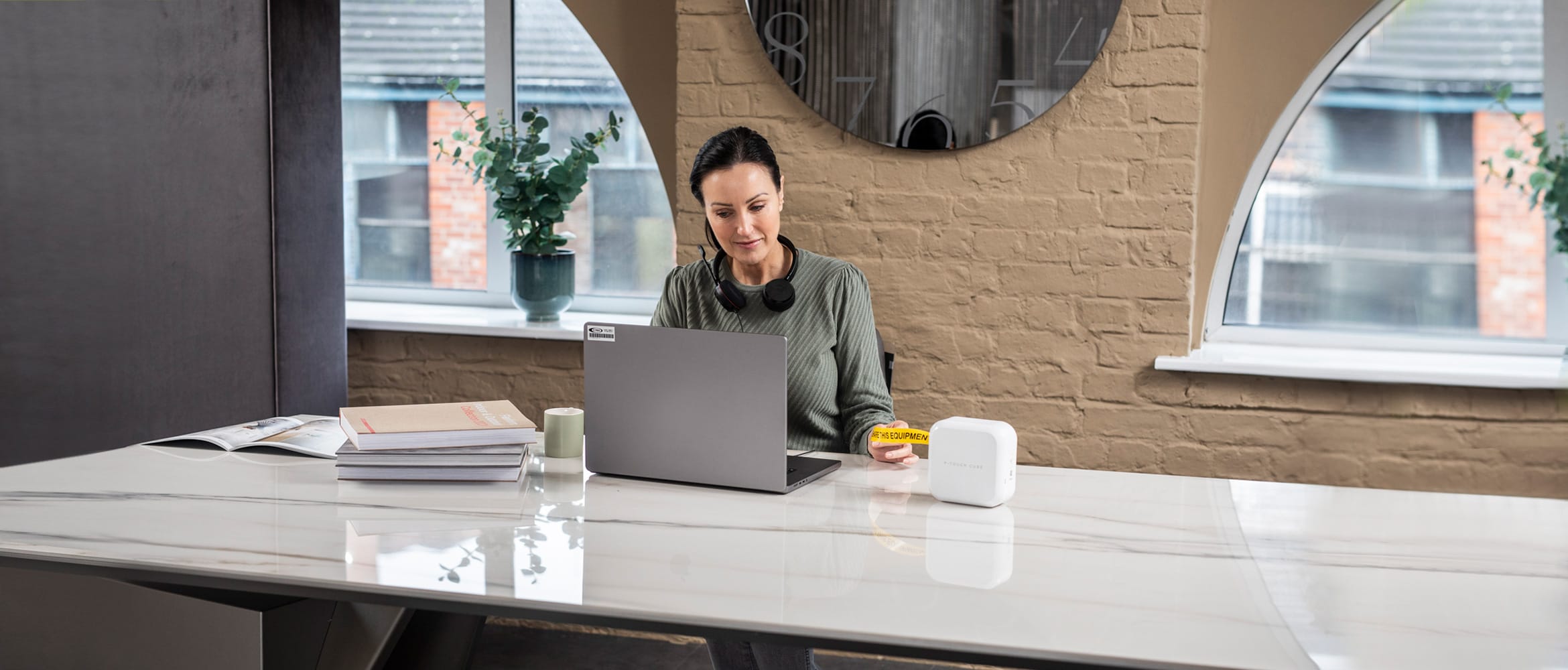 A lady sat at a large white table in front of two half-moon windows is removing a yellow and black label from a Brother label printer which she printed from a notebook computer