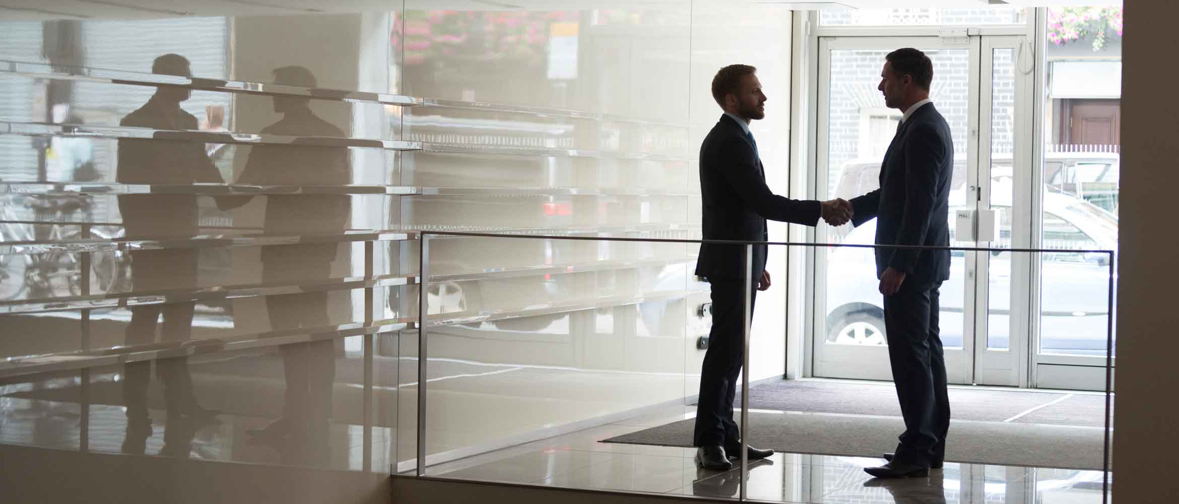 Silhouette of two businessmen shaking hands in office lobby. Male business professional executive colleagues standing near doorway of modern building, handshake.