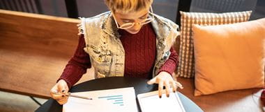 Female freelancer managing finances by comparing printed charts against digital figures on a tablet device