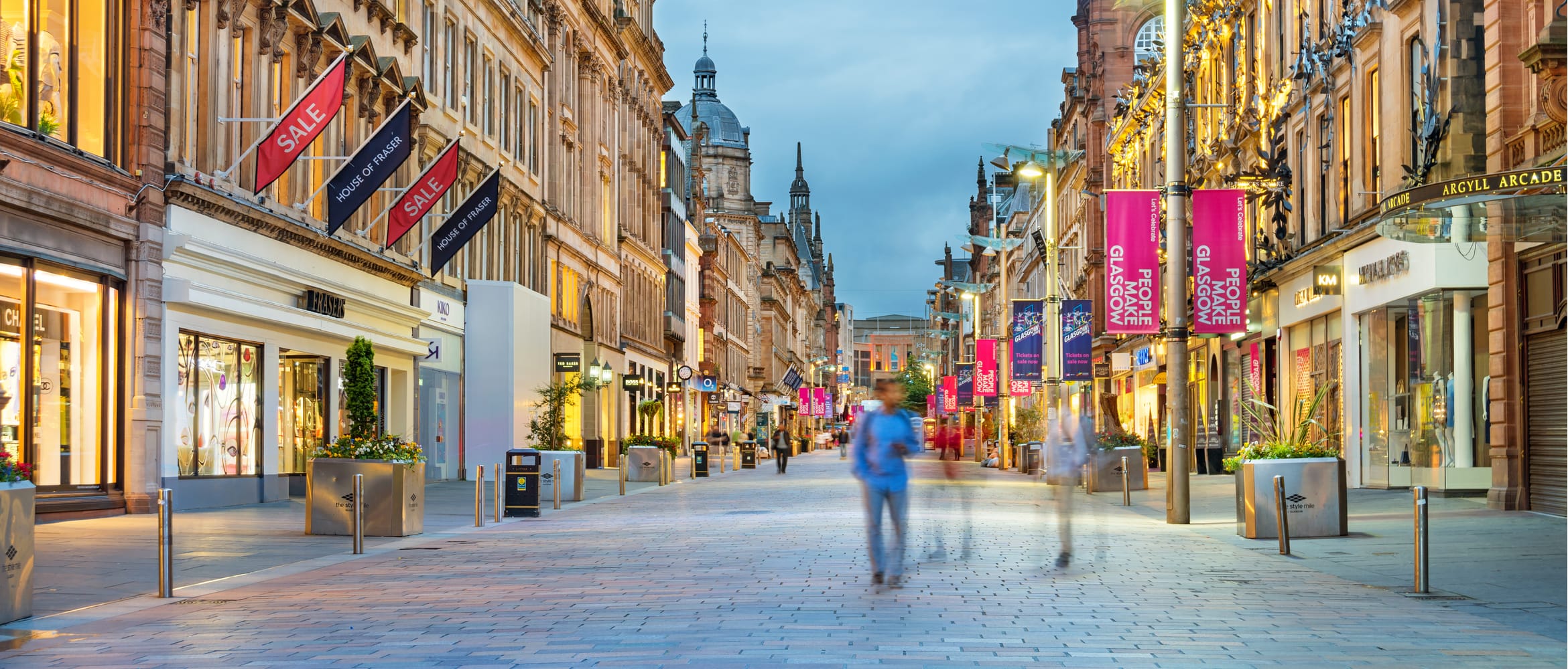 People walking along an almost deserted, pedestrianised, shop lined high street at dusk