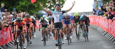 Cyclist Dean Downing with his arms in the air as he crosses the finishing line with spectators behind barriers on both sides of the road and other riders in the background