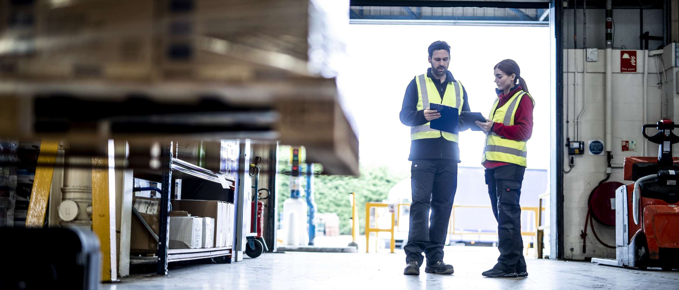 Two workers (one male, one female) are standing in a factory reviewing a document in this transport and logistics warehouse scene