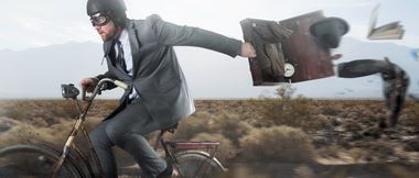 man in a suit and helmet rides a bike while his briefcase blows open