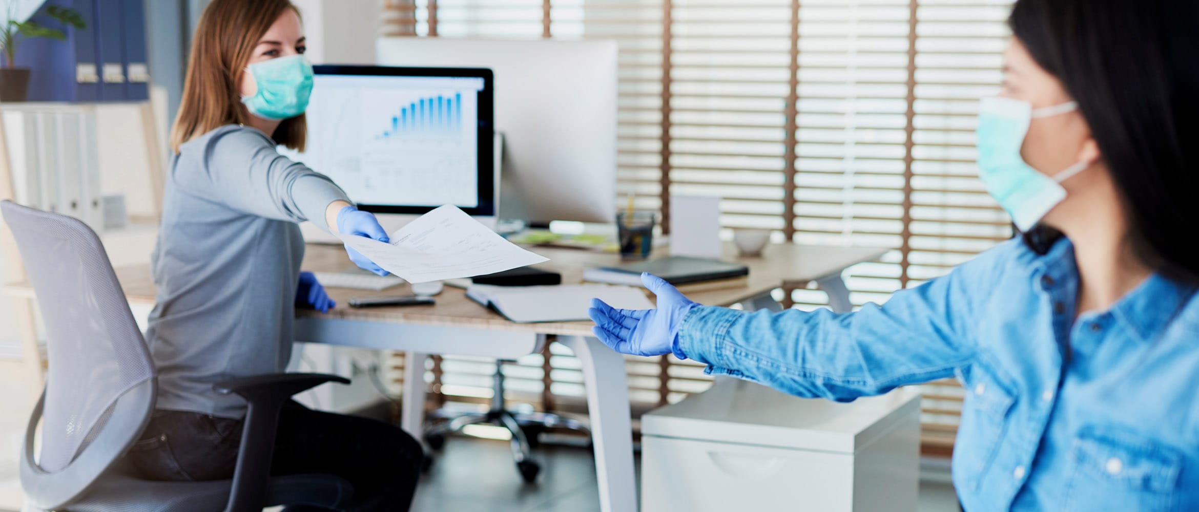 Business woman sat at desk, passing document to female colleague while wearing face covering and surgical gloves in a post-lockdown office environment