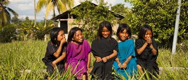 Five young girls smiling while sitting on a sleeper in long grass with palm trees, greenery and a building in the background