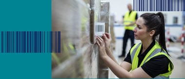 A female warehouse worker wearing a high visibility vest sticking a barcode label to a shrink-wrapped pallet of brown cardboard boxes