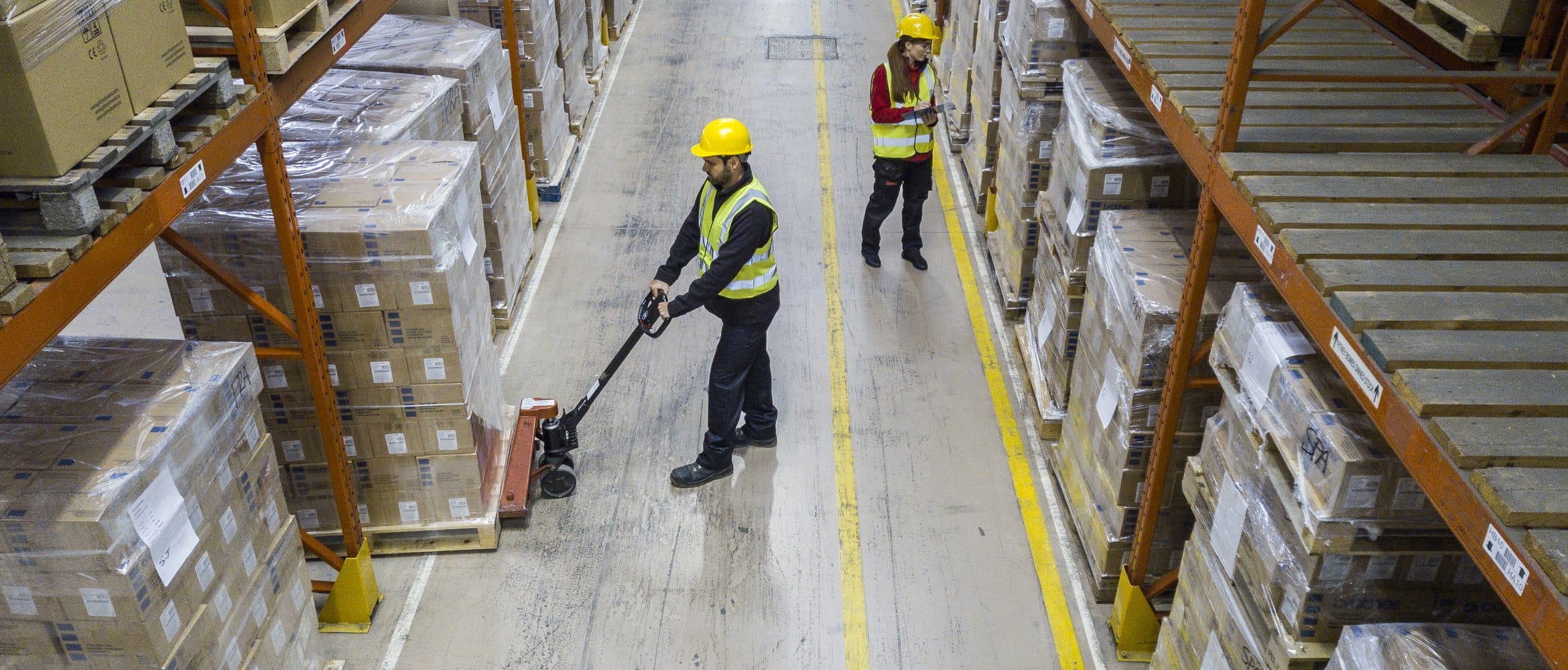 Two warehouse workers inspecting and moving shrink wrapped stock on palettes while wearing hard hats and high vis clothing