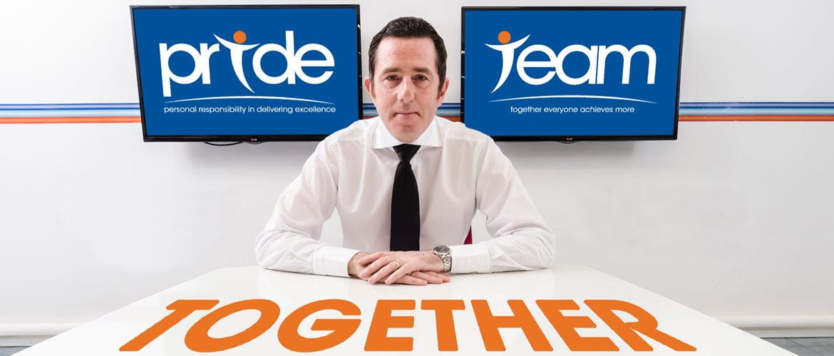 Phil Jones MBE sat at a white table with 'together' written in orange in front of him and two screens behind, one displaying 'pride' and the other displaying 'team'.