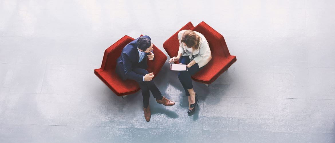 Overhead view of two business people looking at information on a tablet device while sitting on red chairs in a lobby