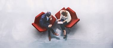 Overhead view of two business people looking at information on a tablet device while sitting on red chairs in a lobby