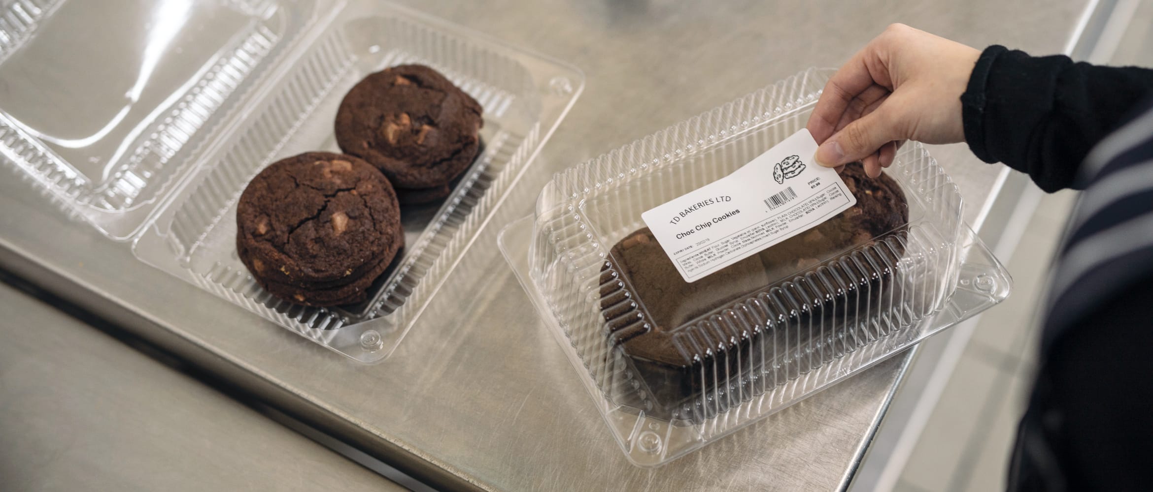 A baker applying an adhesive food description label to a clear plastic box containing choc chip cookies in a kitchen environment