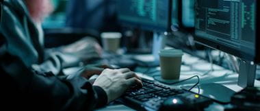 Close-up Shot of Hacker using Keyboard. There is Coffee Cups and Computer Monitors with Various Information