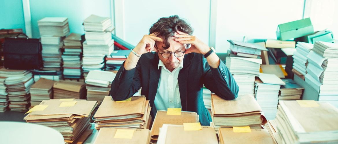 A lawyer holding his head while sat at a desk in an office, surrounded by piles of folders and documents