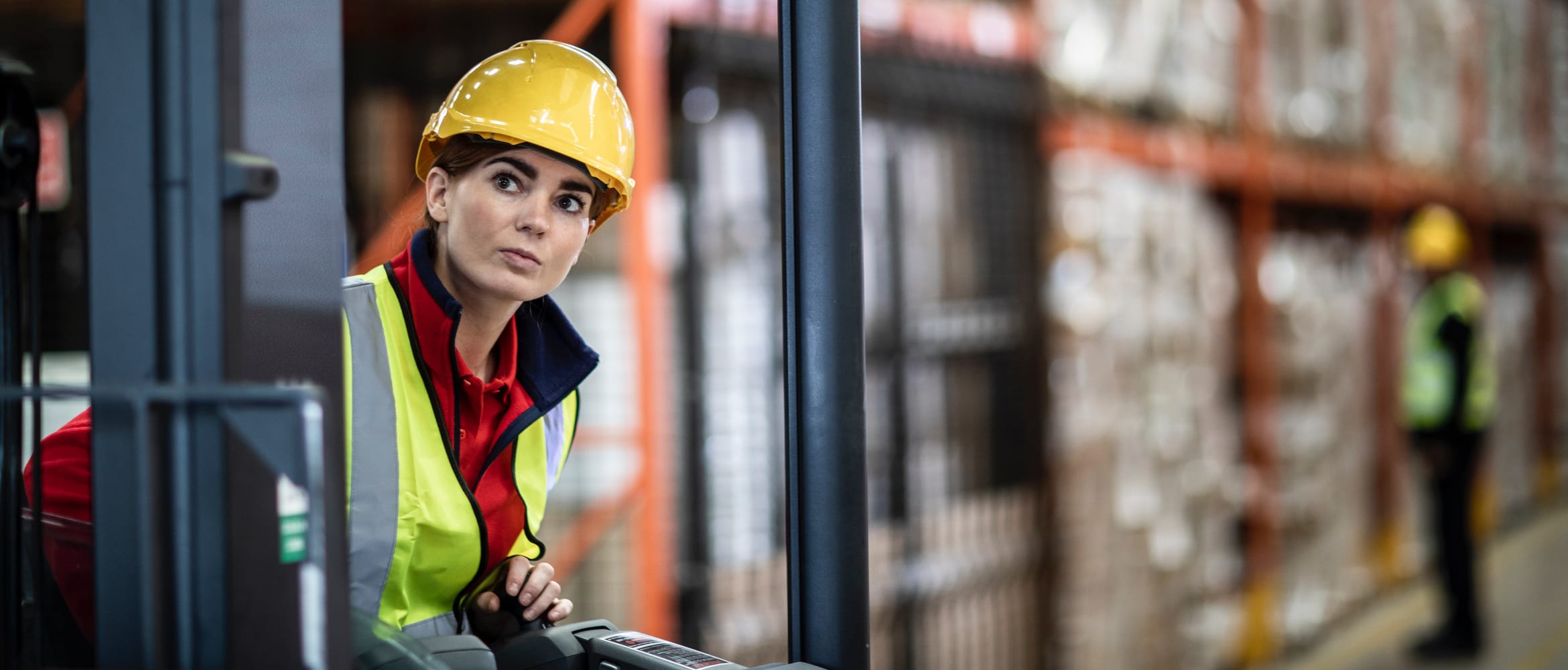 A female warehouse worker wearing a yellow hard hat and high visibility vest while operating equipment with a male colleague inspecting stock in the background