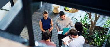 Five employees (two female and three male) in the workplace of the future are holding an informal stand-up business meeting in the office. The scene is pictured from overhead. The team if co-workers are holding files, notepads and tablet computers as they collaborate. 