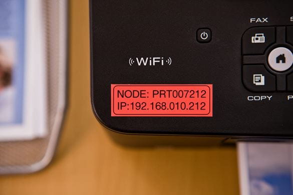 A black on red asset label with node and IP address information stuck to a fax machine
