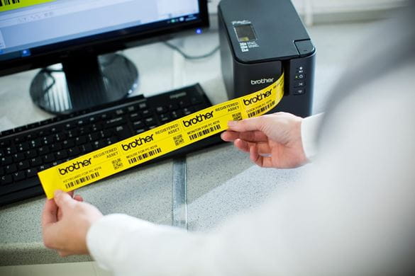 A lady holding a long yellow laminated tape with black asset information and QR codes as it is being printed from a Brother label printer