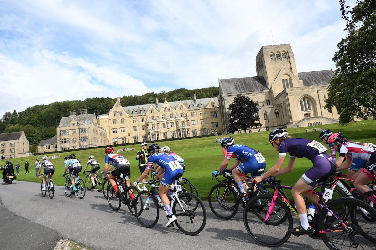 A peloton of female racing cyclists pacing a large, sandstone abbey