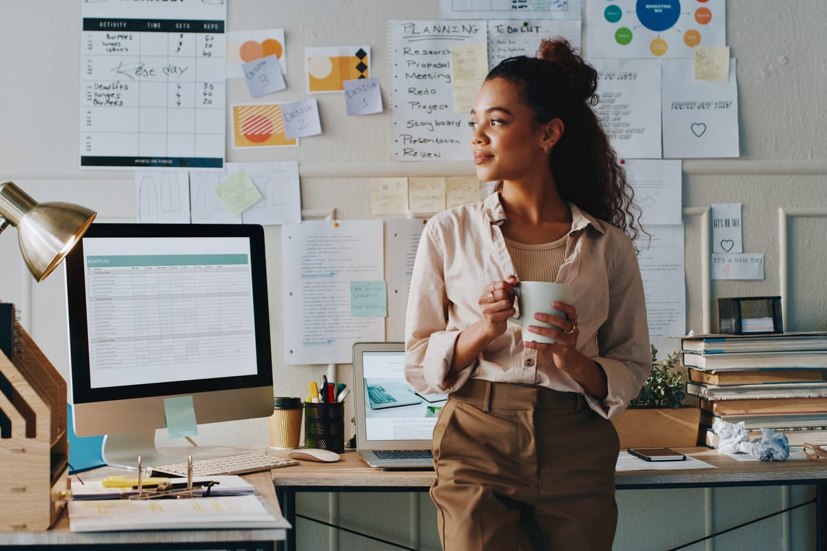 A woman holding a mug, looking contemplative while stood in front of her desk in a home office environment