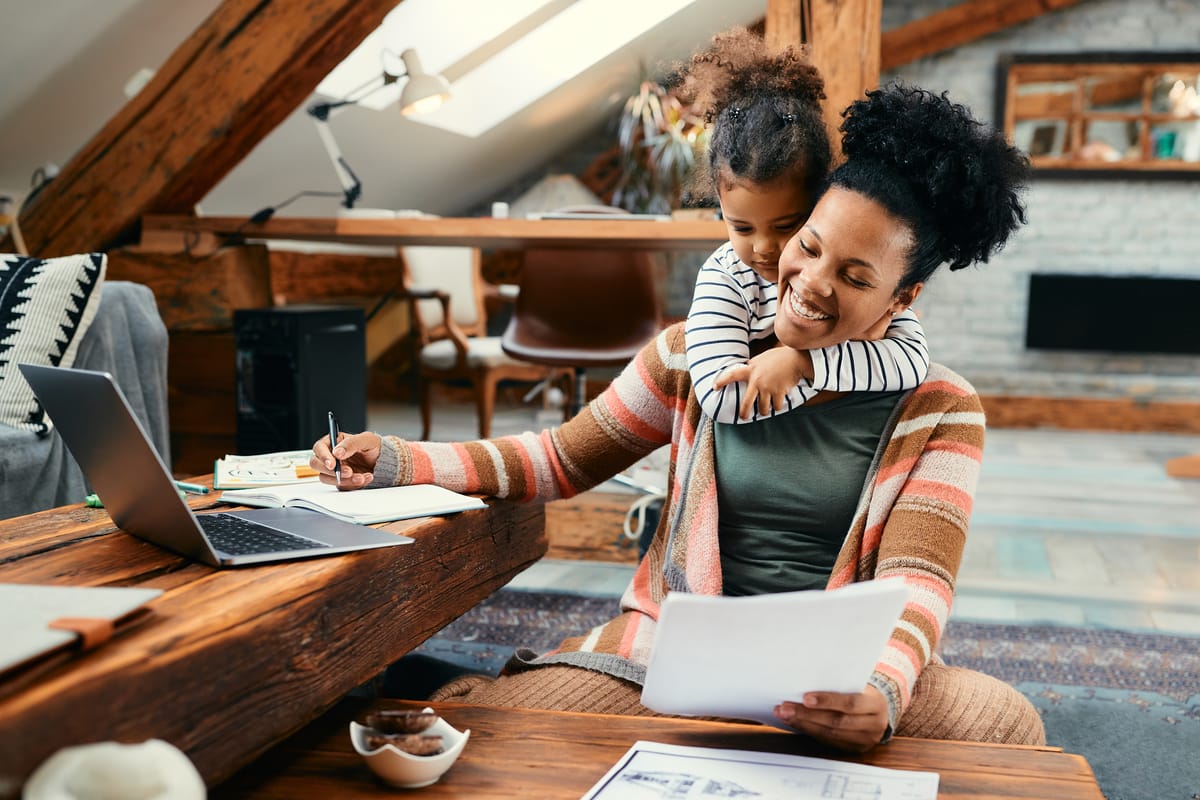 A young girl hugging her busy mother from behind, who is working in a home office loft space