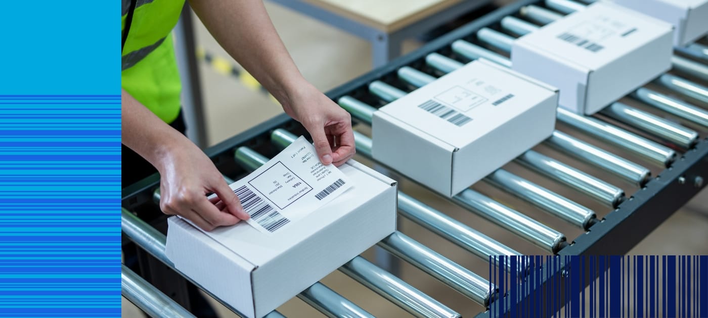 A warehouse worker applying a barcode label to a white cardboard box which is on a roller conveyer