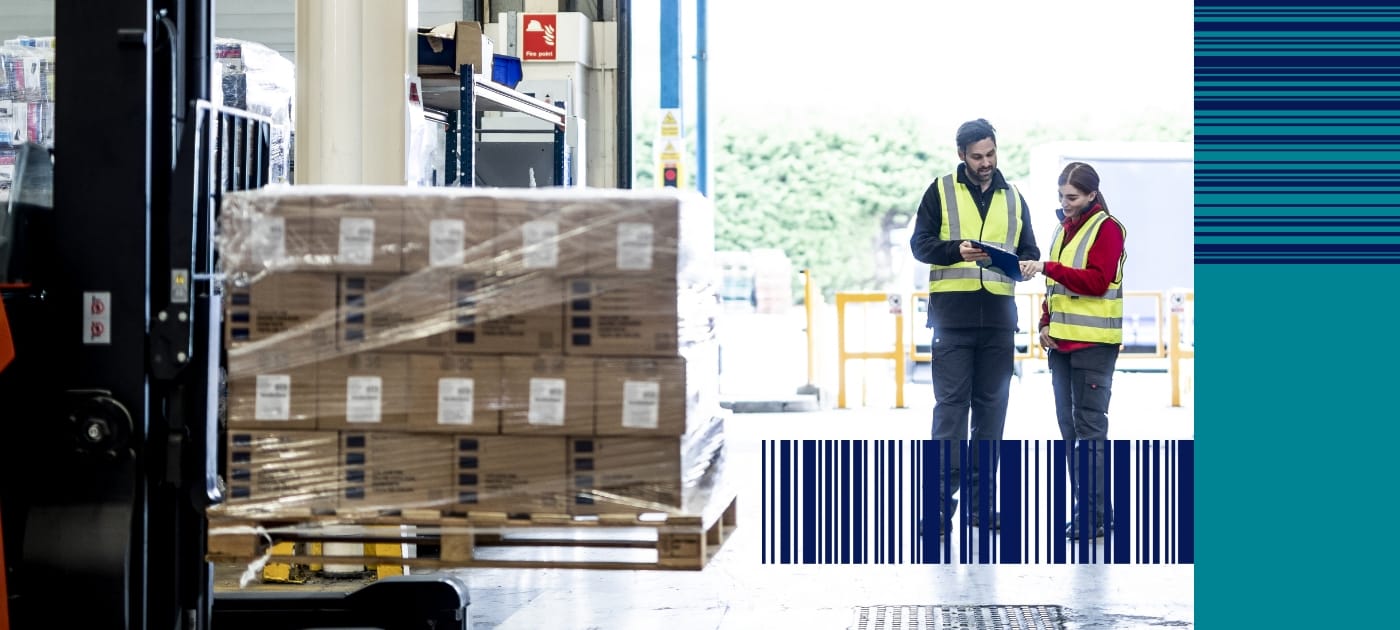 Male and female warehouse workers wearing high visibility vests are looking at a clipboard, a forklift vehicle with a pallet of brown cardboard boxes is in the foreground