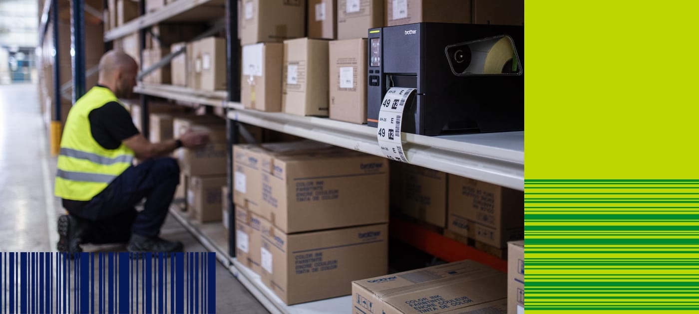 A male warehouse worker wearing a high visibility vest placing brown cardboard boxes on shelves with a Brother industrial label printer in the foreground