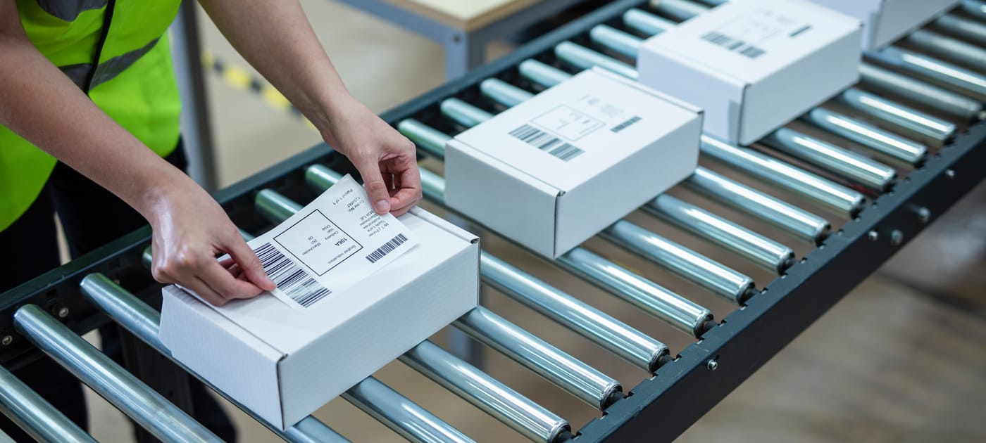 A warehouse worker applying a printed packaging label to a white cardboard box on a roller conveyor system