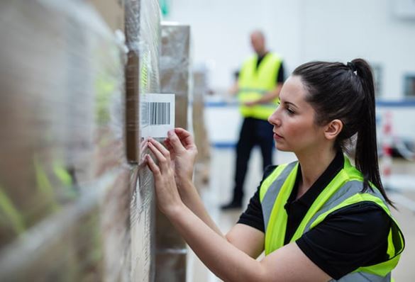 A female warehouse worker applying a barcode label to a stack of shrink wrapped cardboard boxes with a male colleague in the background