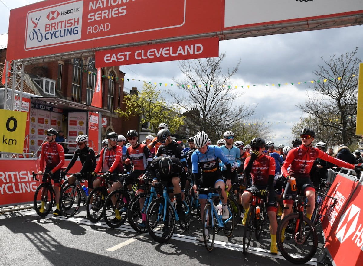 Male racing cyclists, stood with bikes on start line, under start-finish gantry