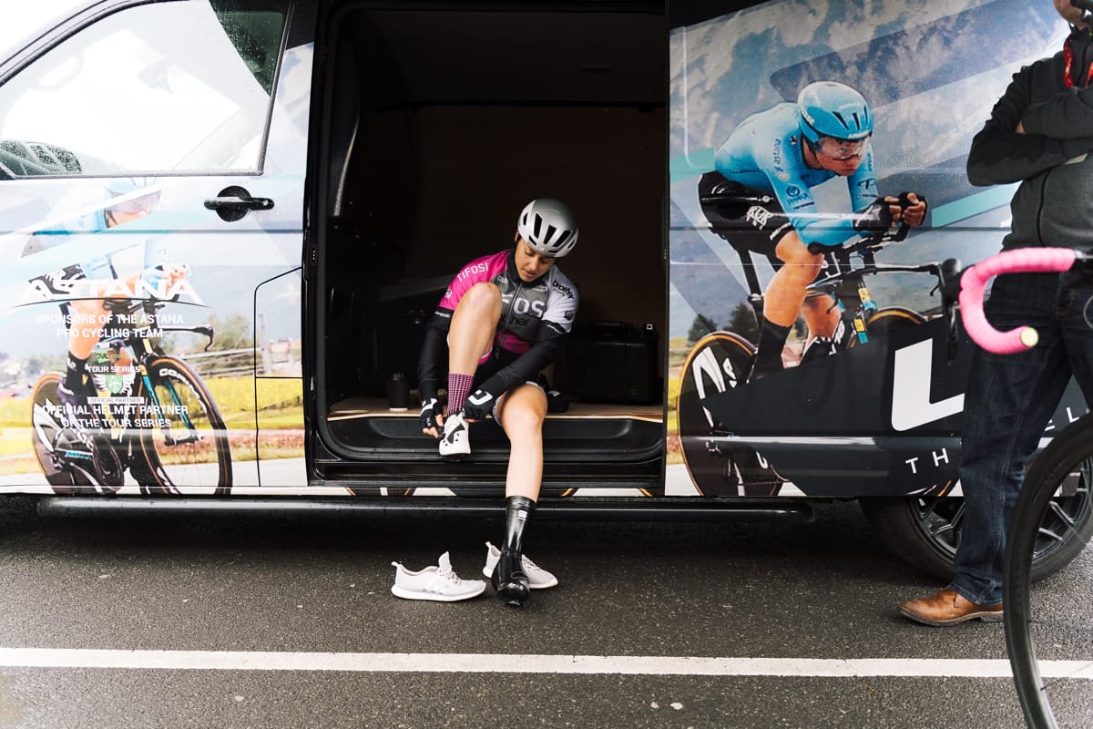 Rebecca Durrell putting on cycling shoes while sitting on the side door sill of a support vehicle