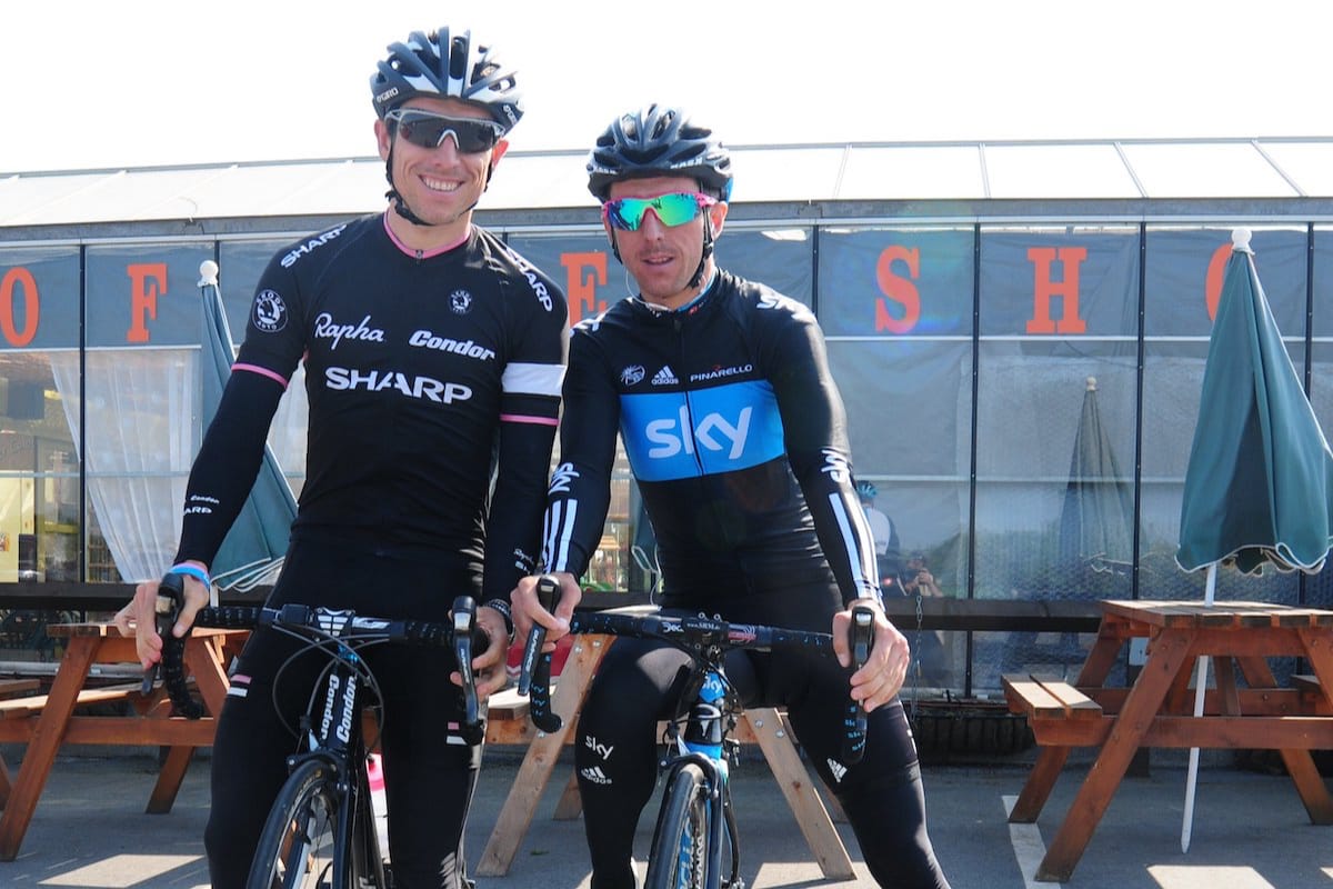 Brothers Dean and Russell Downing wearing cycling clothing while straddling their bikes in front of a glass walled building with tables and parasols in the background