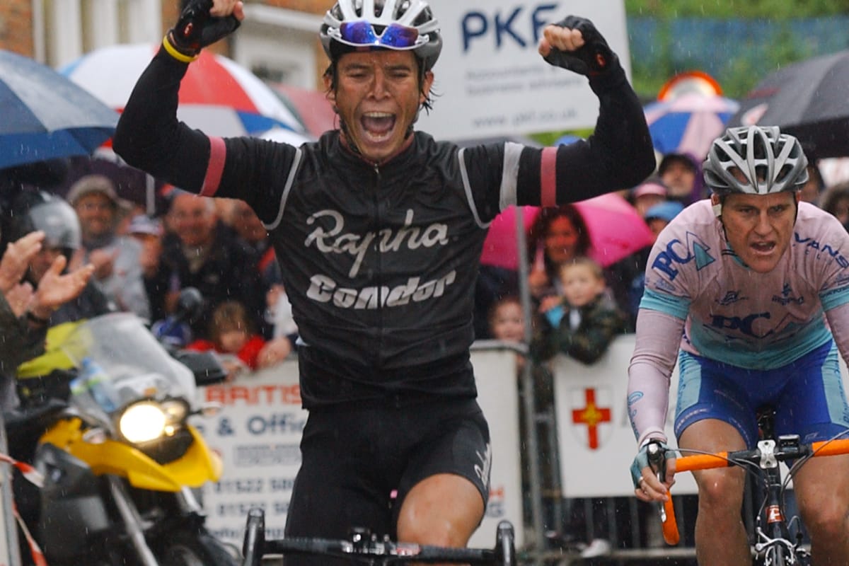 Cyclist Dean Downing with his arms in the air as he crosses the finishing line with spectators and other riders in the background