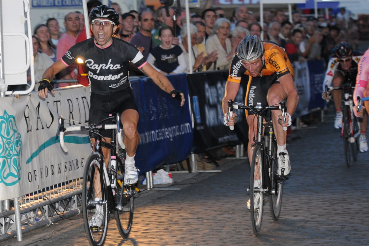 Cyclist Dean Downing crossing the finishing line with another cyclist close behind and spectators cheering behind a barrier